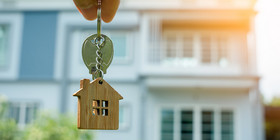 Person hält Wohnungsschlüssel in der Hand und Mehrfamilienhaus im Hintergrund